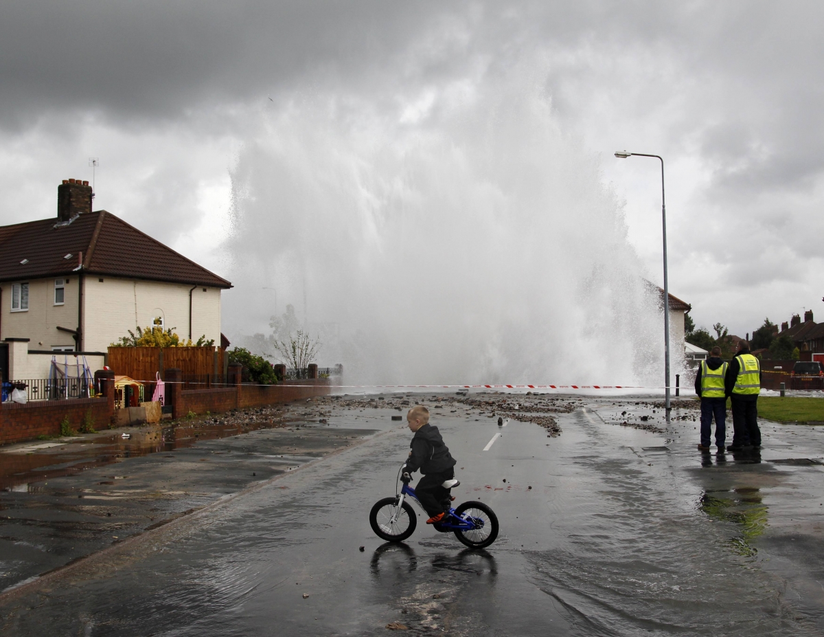 A boy cycles through a puddle as a burst main shoots a plume of water into the air in the Huyton area of Liverpool, northern England, July 18, 2011. The burst main led to localized flooding and evacuations of residents. REUTERS/Phil Noble (BRITAIN - Tags: